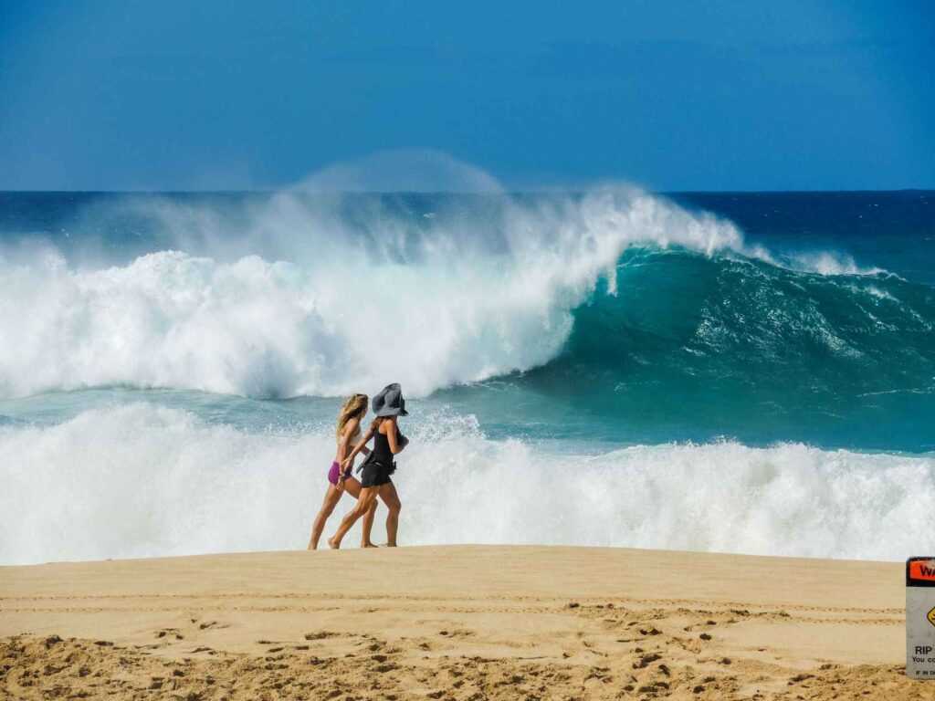 two women walking on sand with huge waves in the ocean during daytime