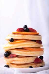 Stack of fluffy pancakes on a plate with red and black fruits scattered around