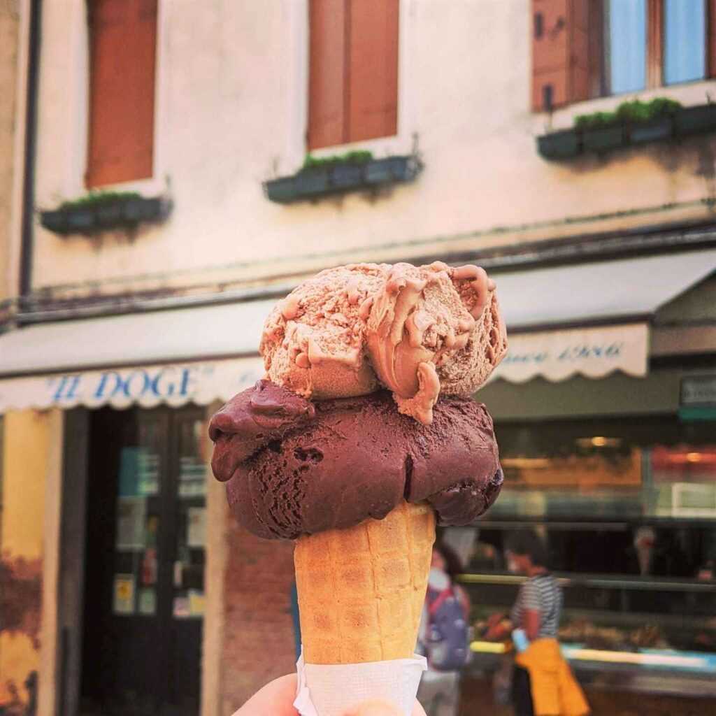 icecream cone with two scoops of icecream being held in front of a shop during day time