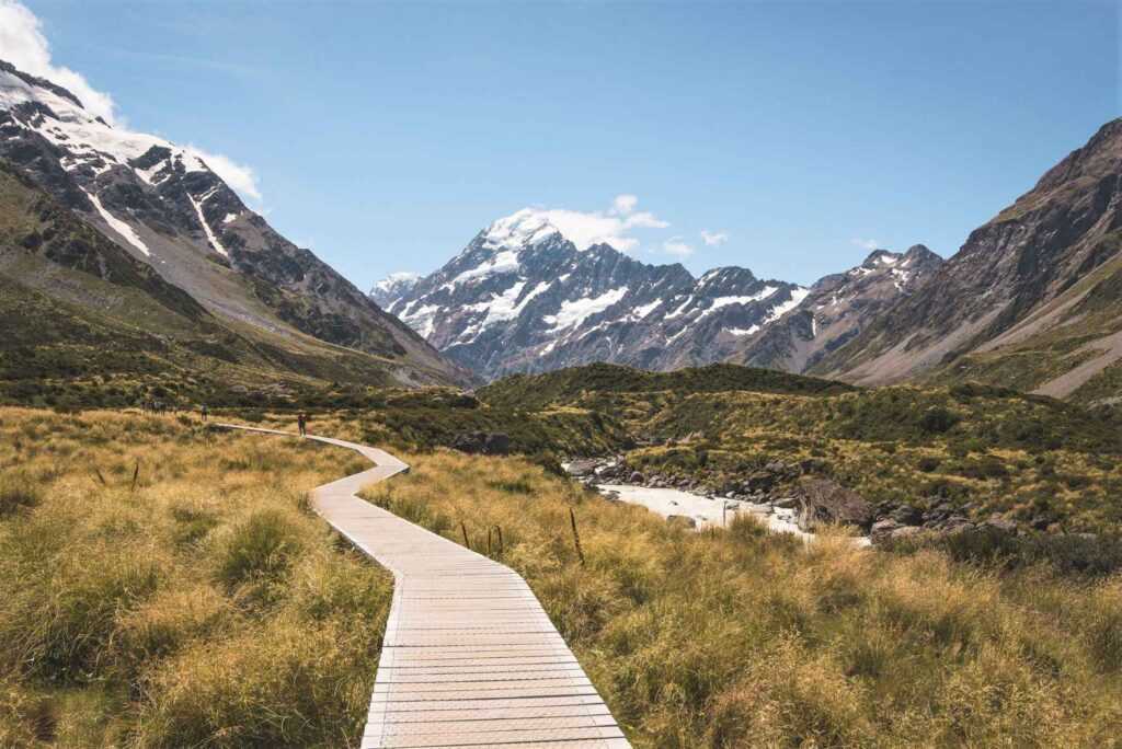 a pathway leading to mountains with grass on either side during daytime. One of the top attractions in New Zealand