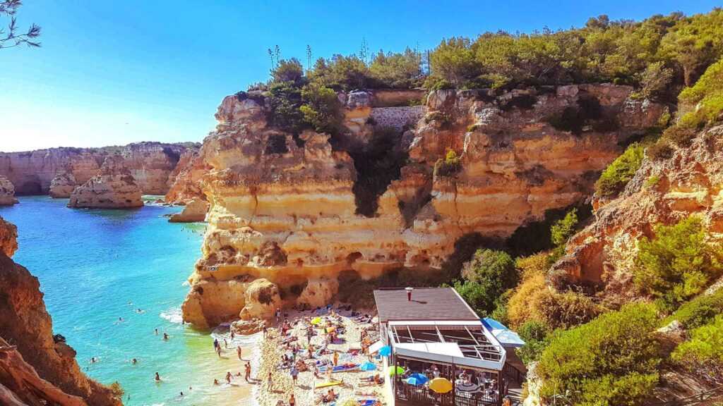 cliffs near a body of water and sand with people sunbathing on the sand during daytime