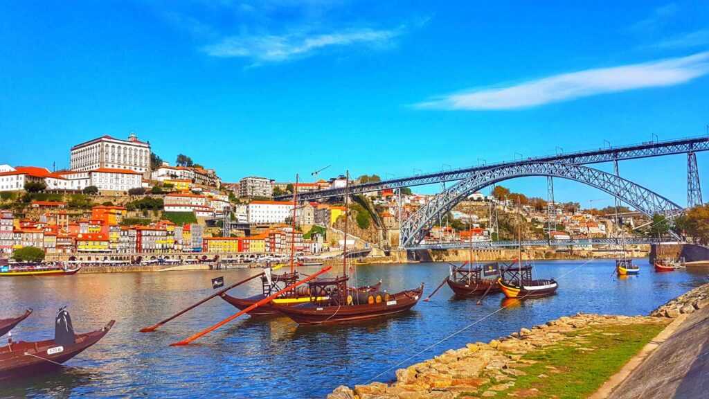 Wooden boats sitting on a body of water with a huge bridge in the background and some colorful houses during daytime. One of the best things to do in Porto 