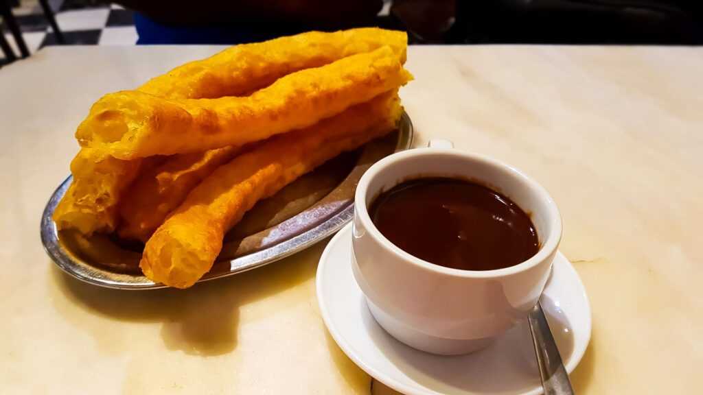 Deep fried dough sticks near a white cup filled with thick chocolate