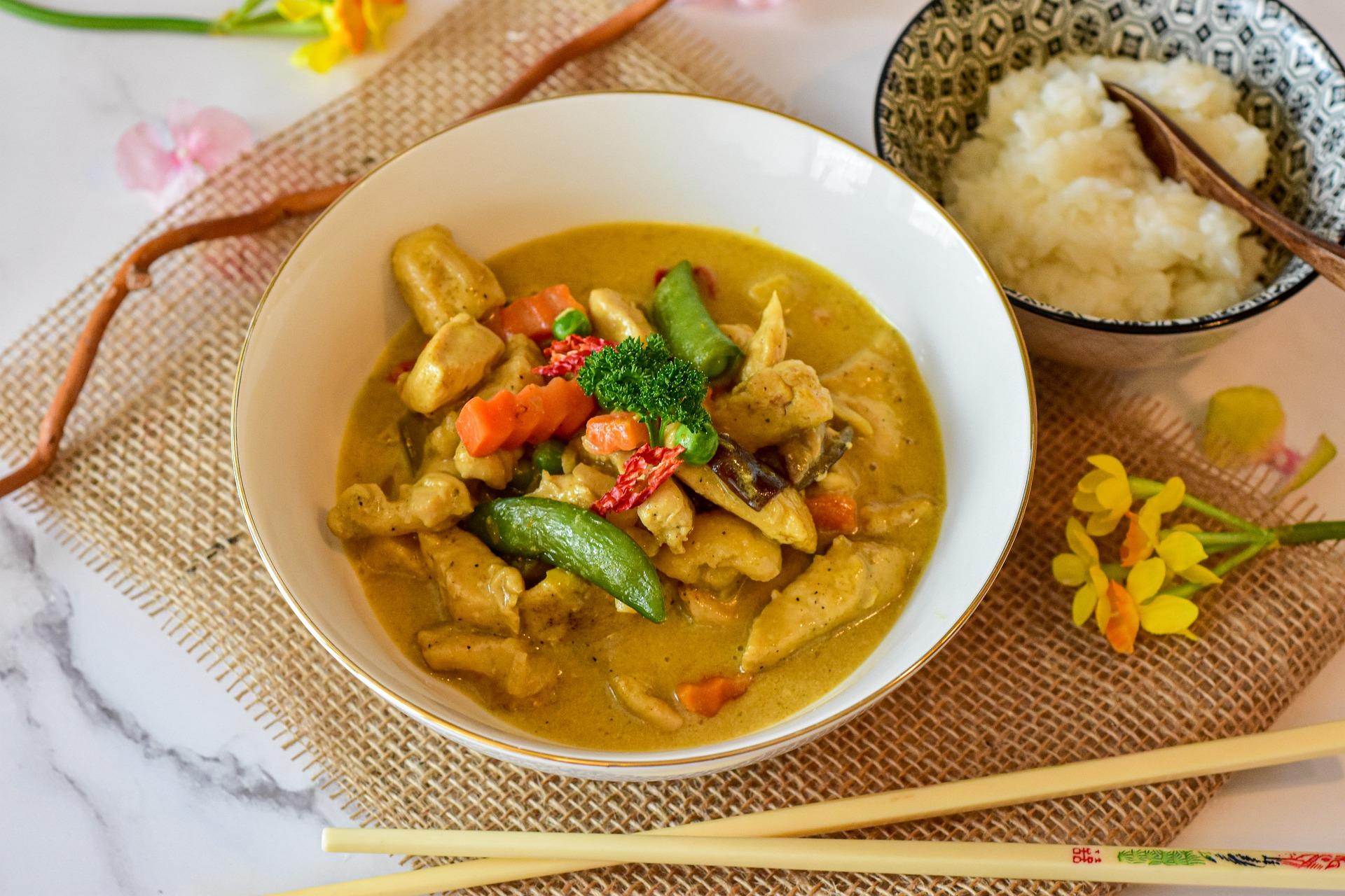 white bowl with yellow curry and vegetables with a bowl of rice and chopsticks beside it