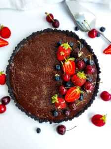 Dark chocolate date topped with red strawberries and cherries on a white table scattered with red berries and a serving spoon