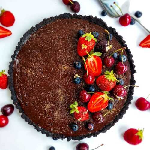 Dark chocolate date topped with red strawberries and cherries on a white table scattered with red berries and a serving spoon