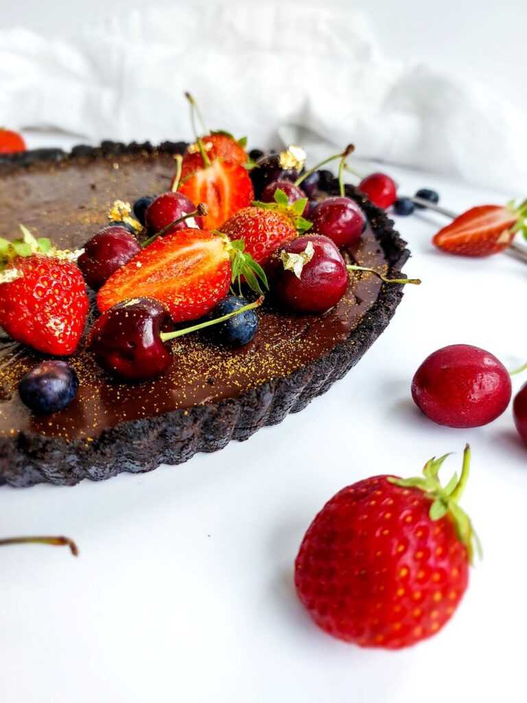 Dark chocolate tart topped with red strawberries and cherries on a white table scattered with red berries