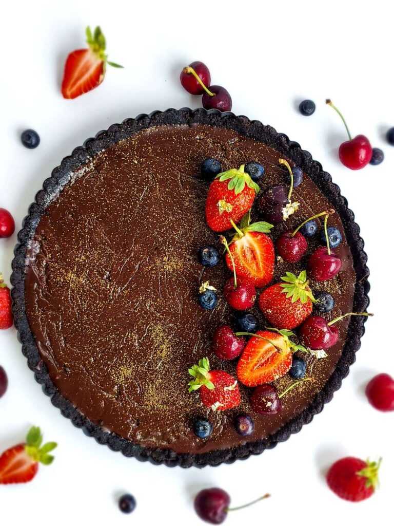 Dark chocolate tart topped with red strawberries and cherries on a white table scattered with red berries 