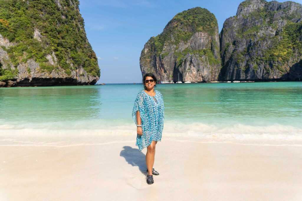 Girl standing in Blue dress on white sand infront of green and blue waters surrounded by limestone cliffs during daytime at the famous Maya Bay