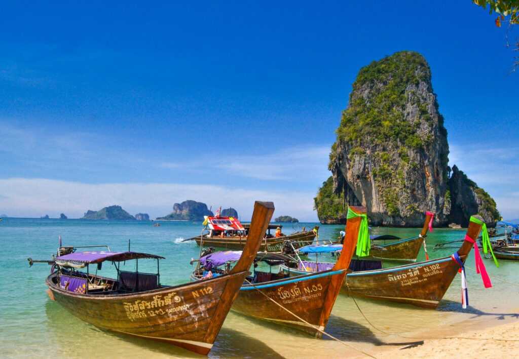 long wooden boats sitting on top of blue water infront of a brown rock covered with green trees during day time