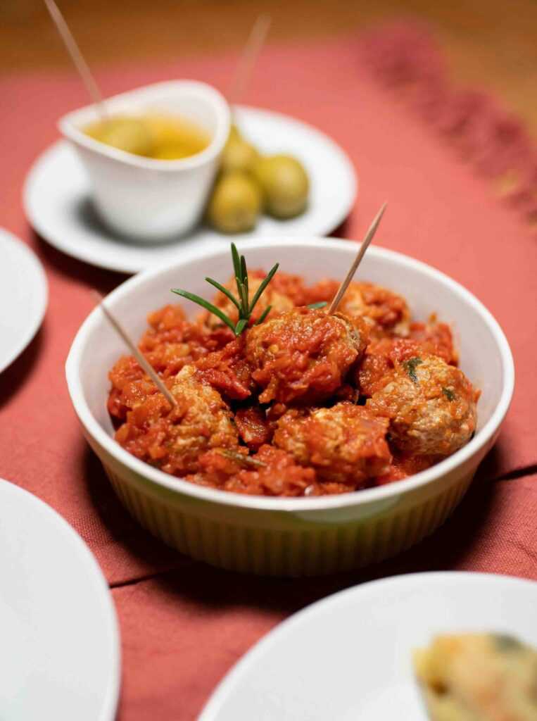 bowl with meatballs with red tomato sauce on a table alongside some green olives