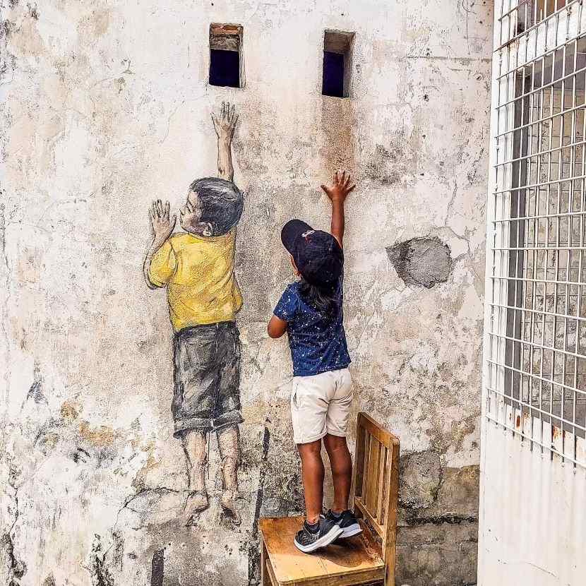 Boy standing on chair with had stretched out next to painting of boy standing on chair